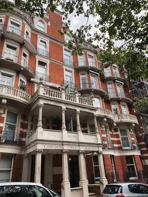 Photograph of a multi-story residential building with a red brick facade and ornate white stone balcony railings. The building features curved bay windows with white trim, and decorative columns supporting the entrance porch marked with the address '43-36' and the name 'Campden Hill Court'. Above, leafy tree branches partially cover the upper floors, casting dappled sunlight on the facade. The ground courtyard area is visible, with parked cars and a clean, well-maintained appearance, embodying the classic architecture typical of South Kensington. The scene is taken during daytime with natural light illuminating the building’s detailed exterior, which appears polished and tidy, fitting within the context of professional cleaning and maintenance services provided by Cleaners South Kensington.
