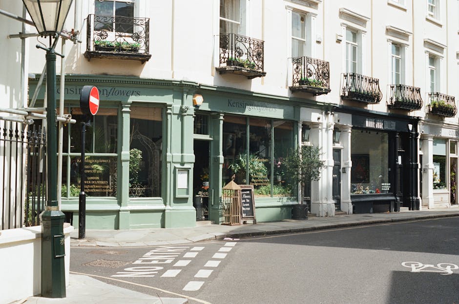 Exterior view of a row of quaint shops on a quiet street in South Kensington, London. The building features white-painted facades with black iron balcony railings on the second floor and large display windows on the ground level. The shop in the forefront, painted in a soft pastel green, displays various plants and flowers inside, visible through the glass. A small blackboard sign outside advertises floral arrangements, and potted plants sit on the sidewalk. The scene is illuminated by natural daylight, highlighting the clean, well-maintained surfaces of the shopfronts. Elements such as the street signage, crosswalk markings, and a bicycle lane are also visible, emphasizing the tidy and organized urban environment, which Cleaners South Kensington ensures remains hygienic and presentable through professional cleaning and sanitisation services.