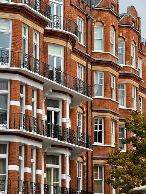 Photograph of a multi-storey red brick residential building in South Kensington, featuring white decorative trim and black wrought-iron balconies on each floor. The windows are white-framed, with some open and others closed, reflecting natural daylight. The building's exterior appears well-maintained, with clean brickwork and tidy balconies. In the lower right corner, part of a tree with green leaves is visible, adding a touch of nature to the urban scene. This image is used in the context of the carpet cleaning case study for South Kensington flat, showcasing the importance of thorough surface cleaning and maintenance, as highlighted by Cleaners South Kensington's professional domestic cleaning services.