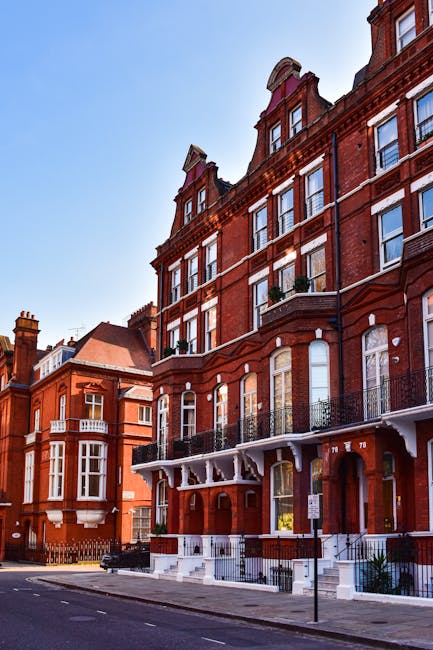 The image shows a row of historic red-brick residential buildings on a clear day, with large sash windows, decorative white trims, and small balconies with black wrought iron railings. The ground-level entrance has steps leading up to white double doors, framed by white arches. Street-level pavement is clean, and the scene is illuminated by natural daylight, with no visible signs of dust or dirt. This exterior shot depicts well-maintained, classic architecture typical of South Kensington, emphasizing the building facades and surrounding street, which is tidy and free of debris. The setting aligns with residential interior or exterior cleaning case studies by Cleaners South Kensington, illustrating neatness and the importance of building maintenance in urban settings.