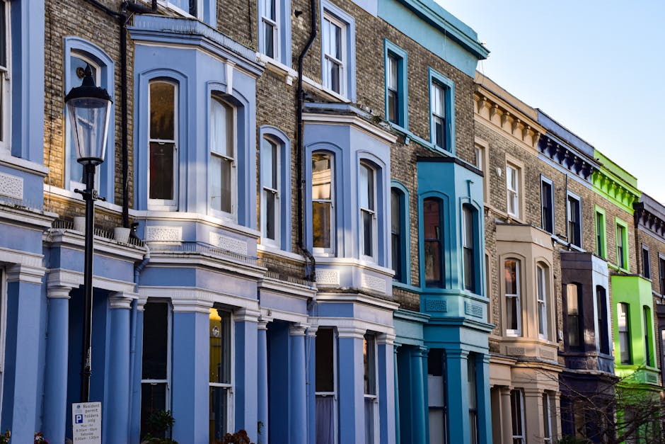 A row of Victorian-style terraced houses with multi-colored facades, including shades of blue, beige, and green, situated along a street in South Kensington. The buildings feature tall, arched windows with white frames, some with decorative molding, and are topped with intricate cornices. A black lamppost is visible in the foreground, and the street appears clean and well-maintained. The natural daylight illuminates the facades, highlighting their architectural details. This scene reflects the elegant residential character typical of South Kensington, where professional cleaning services by Cleaners South Kensington ensure the upkeep and hygiene of such properties, especially useful for end of tenancy cleaning and maintaining domestic cleanliness.
