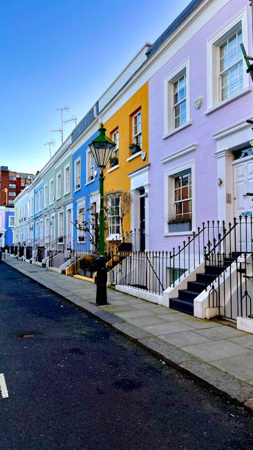 A row of vibrant, pastel-colored terraced houses on a residential street in South Kensington, featuring painted facades in yellow, lavender, and light blue. The houses have white-framed sash windows, black wrought iron railings along the staircases leading to front doors, and black street lamps positioned on the sidewalk. The street is paved with asphalt, and the sidewalk is made of concrete slabs, illuminated by natural daylight. This exterior scene highlights the charming, well-maintained appearance of the neighborhood, illustrating the importance of surface cleaning and maintenance for enhancing property value in line with end of tenancy cleaning tips for landlords, as promoted by Cleaners South Kensington.