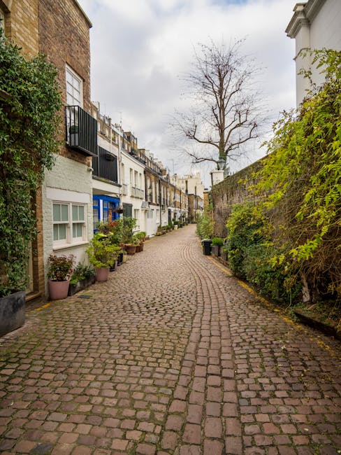 A narrow, cobblestone residential street in South Kensington with a gentle curve leading towards the background. On the left side, there are several small potted plants placed along the base of the white and light-colored terraced houses, which feature large windows, some with white frames, and small balconies with black railings. On the right side, a tall brick wall covered partially by green climbing plants and a neighboring building with a white facade. A tall, leafless tree stands prominently at the center background, with overcast sky above. The scene is clean and well-maintained, typical of a quiet, historic neighborhood. This image exemplifies surface cleanliness and outdoor maintenance associated with residential standards emphasized by Cleaners South Kensington, aligned with the Kensington and Chelsea council bulky waste cleaning rules.