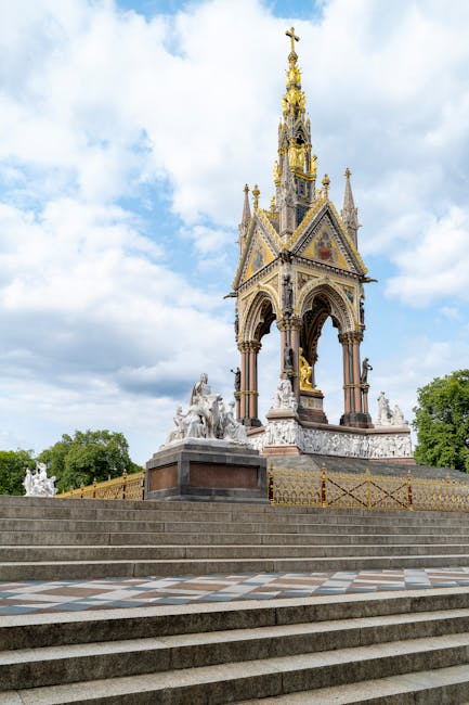 A detailed view of the Victoria Memorial in front of the Victoria and Albert Museum, showing the ornate marble structure with decorative sculptures and gilded accents. The monument features a tall, gi