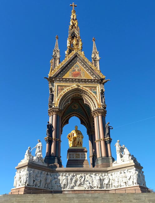 A detailed view of the Victoria Memorial in front of the Victoria and Albert Museum, showing the ornate marble structure with decorative sculptures and gilded accents. The monument features a tall, gi